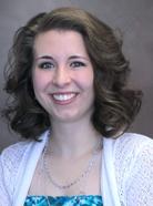 A woman with short, wavy hair smiles at the camera. She is wearing a white cardigan over a blue patterned top and a silver necklace. The background is plain and neutral-colored.